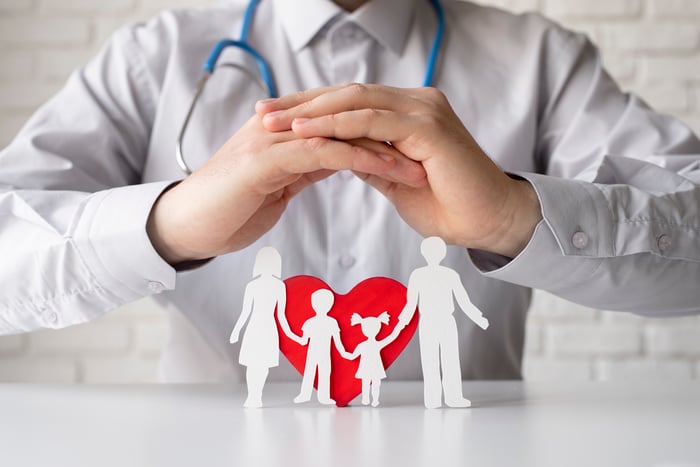 Close-up of a doctor with a stethoscope protecting a paper-cut family and red heart, symbolizing healthcare, safety, and patient-centered care.