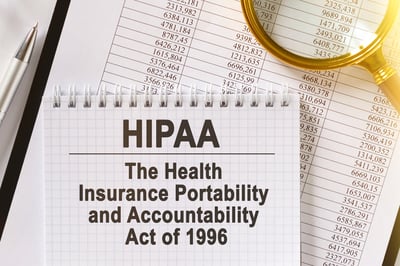 Notebook with the text “HIPAA – The Health Insurance Portability and Accountability Act of 1996” placed on top of financial documents with a magnifying glass and pen.