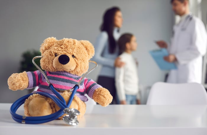  Teddy bear with a stethoscope on a table in a pediatric clinic, with a doctor consulting a child and parent in the background, representing child-friendly care and effective digital marketing strategies for pediatricians.