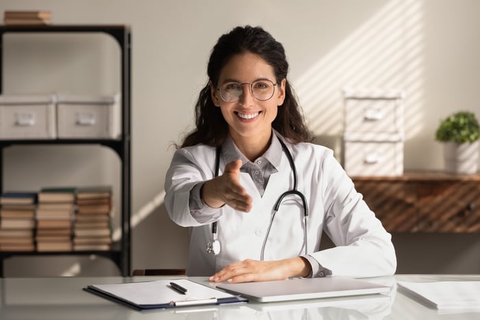 Female doctor in a white coat and glasses smiles while reaching her hand out for a handshake, symbolizing welcoming new patients through an AI-driven patient acquisition engine.