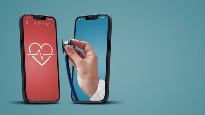 A visual representation of the 2026 telemedicine scope featuring a doctor’s hand extending a stethoscope from one smartphone to another, symbolizing the seamless delivery of remote clinical services and virtual heart health monitoring.