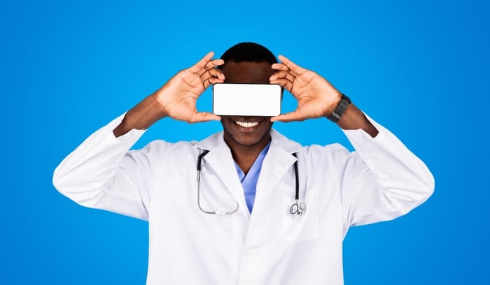 Smiling doctor holding a smartphone over his eyes against a blue background, symbolizing the fusion of modern technology with vision care. Represents the role of mobile-friendly, senior-accessible digital tools in ophthalmology practice growth.