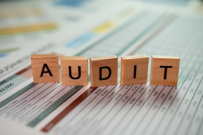Close-up photograph of five wooden scrabble-style tiles spelling out the word "AUDIT" laid across a blurred financial or data spreadsheet containing rows of numbers and figures, symbolizing a review of finances or performance.