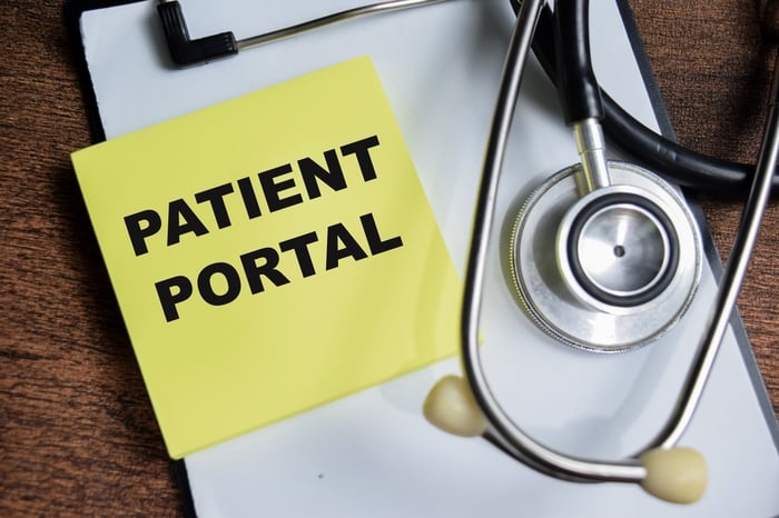 Close-up of a stethoscope and clipboard with a sticky note labeled 'PATIENT PORTAL', representing the digital solution for parent self-service and reducing clinic overload.
