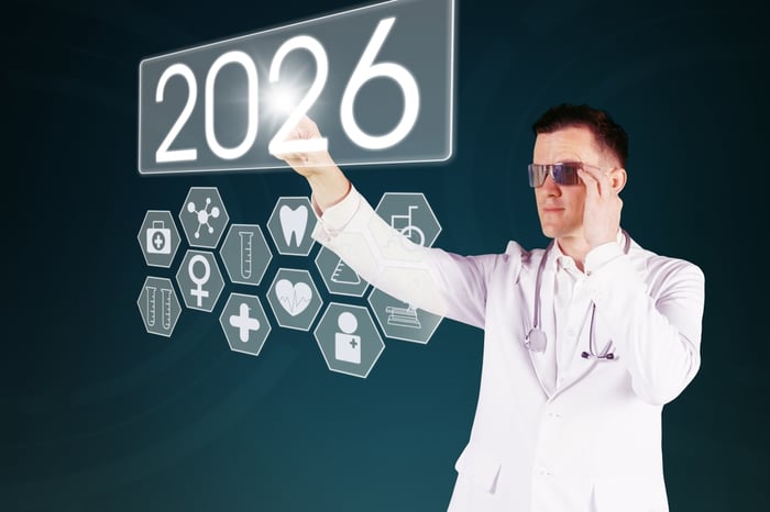 A male medical professional in a white coat and futuristic glasses interacting with a glowing digital touch interface displaying '2026' alongside holographic medical icons for dentistry, cardiology, and pharmacy.
