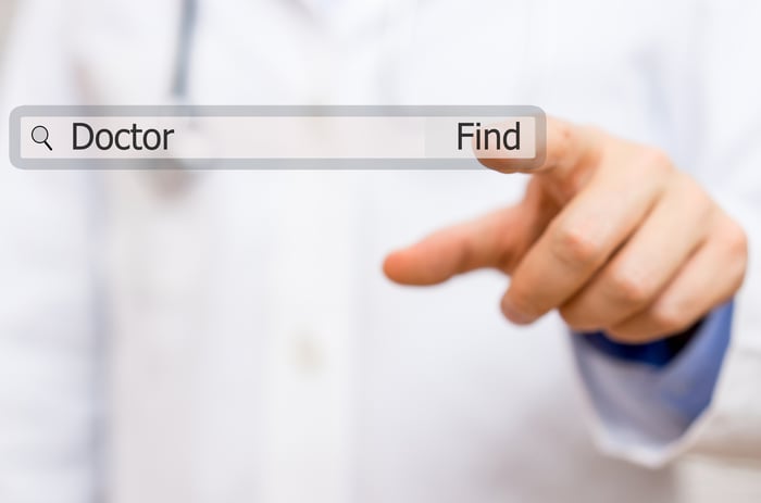 Hand in doctor's coat clicking 'Find' on a search bar with the query 'Doctor' for online doctor search and appointment book