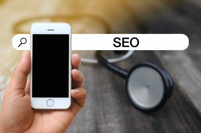 Close-up of a hand holding a smartphone with a stethoscope in the background and the word "SEO" in a search bar — symbolizing the importance of local search optimization in healthcare and dermatology practices.